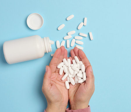 A woman's hand holding a plastic bottle and white oval pills on a blue background, top view.の写真素材