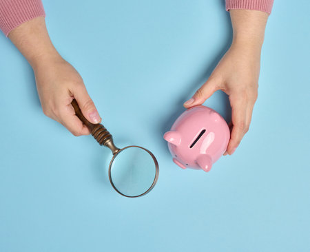 A woman's hand holds a piggy bank and a magnifying glass on a blue background, top viewの写真素材