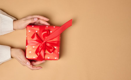 Female hands holding a gift box with a red ribbon on a brown background. View from aboveの写真素材