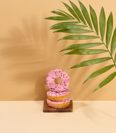 Donut covered with pink glaze and sprinkled with multi-colored sprinkles, brown background with a shadow from a palm leafの写真素材