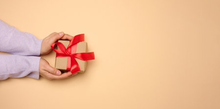 A woman's hand holds a gift box wrapped in a red silk ribbon on a beige background, top view. Copy spaceの写真素材
