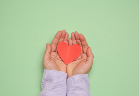 A woman's hand holds a red paper heart on a green background, symbolizing the concept of donation and loveの写真素材