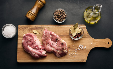 Two raw pork neck steaks on a board and spices for cooking. Top view of black tableの写真素材