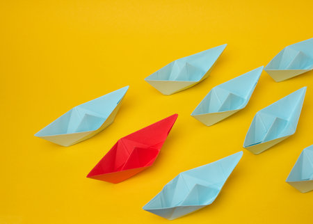 Group of blue paper boats follow red against a yellow background. Strong leader concept, mass manipulation. Starting a business with a well-coordinated team, start-upの写真素材