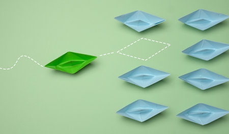 Group of blue paper boats heading in one direction and one green one heading in the opposite...