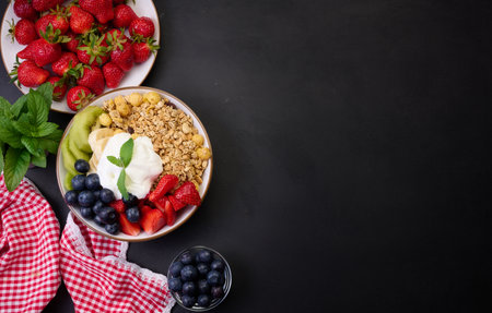 Healthy breakfast. Granola with strawberries, kiwi, banana and blueberries in a round plate on a black table. Healthy and tasty food, copy spaceの写真素材
