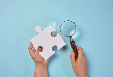 White empty puzzle and a magnifying glass in hand on a blue background, top viewの写真素材