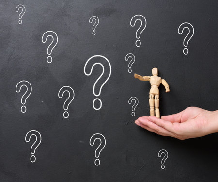 A woman's hand holding a wooden miniature puppet against a black chalkboard with chalk-drawn question marks. Concept of searching for truth, gathering informationの写真素材