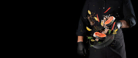 Chef in black uniform holding frying pan and levitating pieces of raw salmon fish and spices on black background. Space for descriptionの写真素材