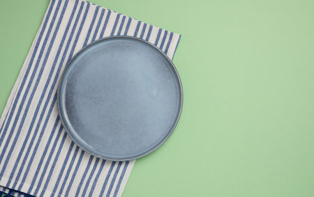 Empty round ceramic plate and blue kitchen towel on green background, top viewの写真素材