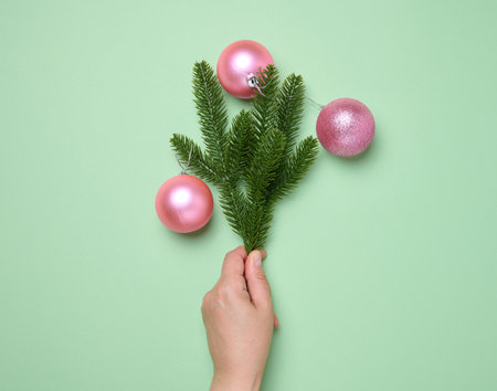 Female hand holding shiny pink balls and green spruce branches on green background, Christmas backgroundの写真素材