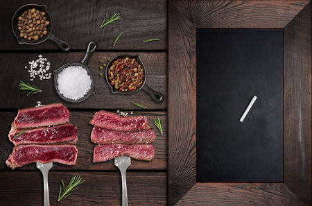 Juicy pieces of roast beef are skewered on a fork, spices. Nearby is an empty black chalk board for writing a menu or recipeの写真素材