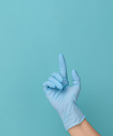 Female hand with blue latex medical glove on blue background, index finger raised upの写真素材