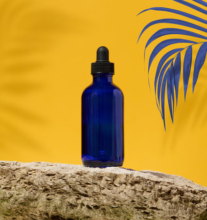 Blue glass bottle with black pipette and palm branch on yellow backgroundの写真素材