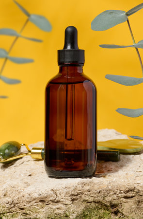 Transparent brown glass bottle with black pipette and eucalyptus branches stands on stone, yellow background. Cosmetics and oilsの写真素材