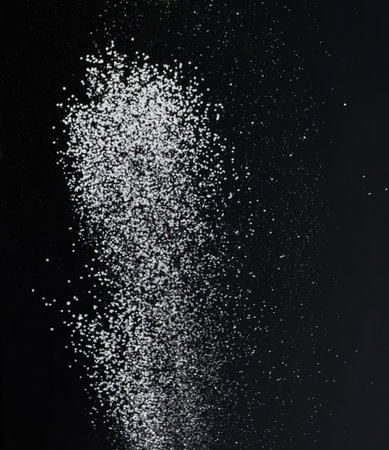 Close-up of white granular particles sugar, falling against a black background. High contrast and detailed texture of the grainsの写真素材
