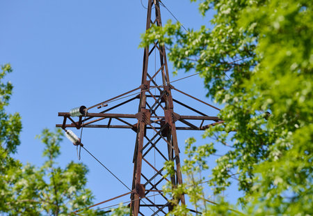 Upper part of an old, rusty power line tower against a blue sky and green tree leaves. Seen from a bottom-up perspective.の写真素材