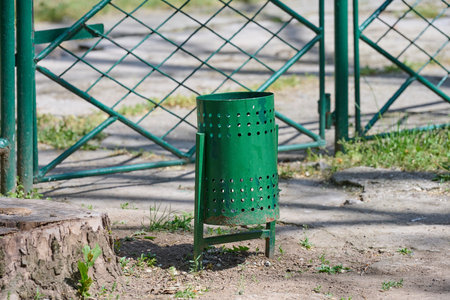 A green perforated metal trash can stand in a park or public area. Outdoor waste disposal binの写真素材