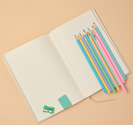 Top view of an open notebook with pastel-colored pencils, green sharpener, and turquoise eraser on a beige background. Flat lay composition of school or office stationery supplies.の写真素材