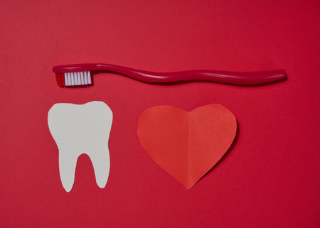 Red toothbrush, white paper tooth cut out and red paper heart placed on red background. Top view.