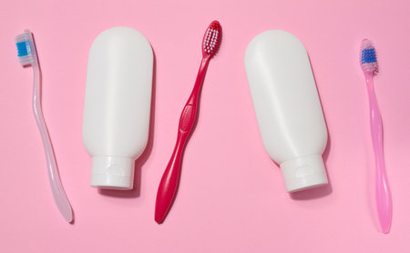 Three toothbrushes and two white plastic tubes of toothpaste placed on a pink background. Top view.