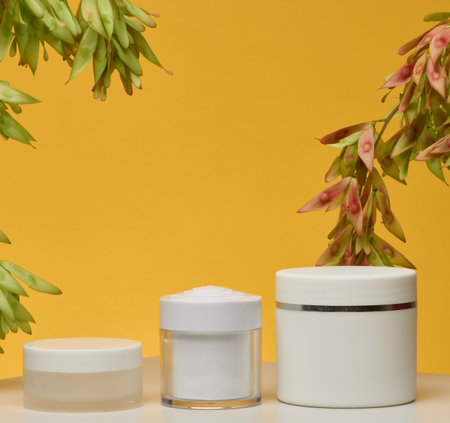 Three white cosmetic jars without labels stand on a white background. Branches with green and reddish-pink seeds hang from above, yellow background.の写真素材