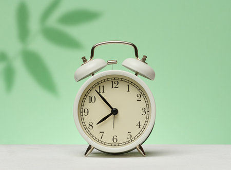 White vintage alarm clock stands on a green surface against a background with shadows from leaves....