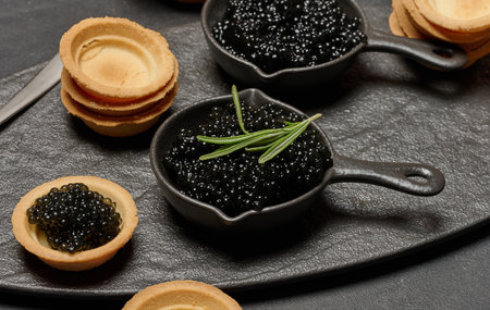 Black caviar in cast iron pans and tartlet on dark board.