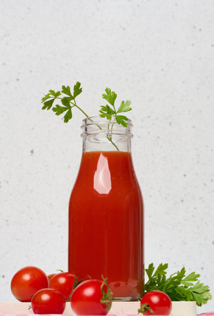 Glass bottle with tomato juice decorated with parsley. Nearby are fresh red tomatoes and parsley leaves on a light background.の写真素材