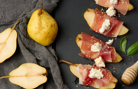A slice of pear wrapped in prosciutto, topped with cheese and sesame seeds on a black slab, top viewの写真素材
