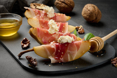 Sliced ââpear wrapped in prosciutto, topped with cream cheese and sesame seeds, served on an elegant black slab plateの写真素材