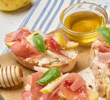 Italian bruschetta with prosciutto, pear, soft cheese, honey and sesame seeds. The dish is served on a light marble boardの写真素材