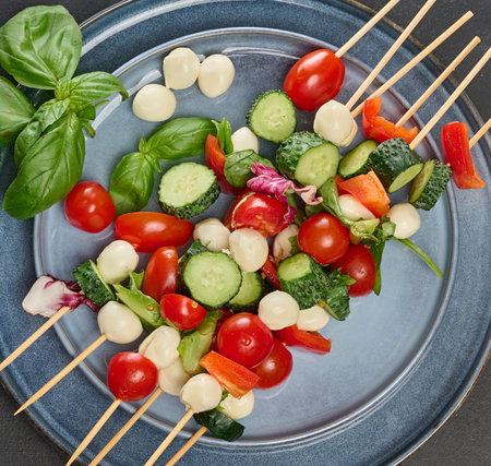 An assortment of vegetable and cheese canapes on wooden skewers served on a blue plate, cherry tomatoes, cucumbers, sweet red peppers, lettuce leaves, mozzarella cheese and fresh basil.の写真素材