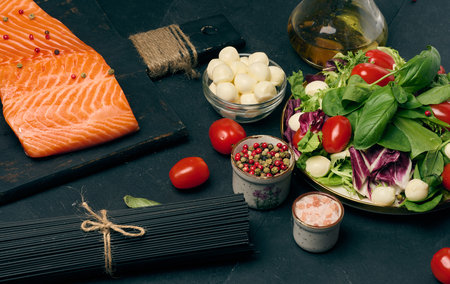 Whole piece of fresh salmon on a black cutting board, next to a bowl of fresh salad, mozzarella and black pasta. Healthy dinner.の写真素材