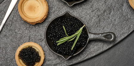 Black caviar in cast iron pans and tartlet on dark board. Top view
