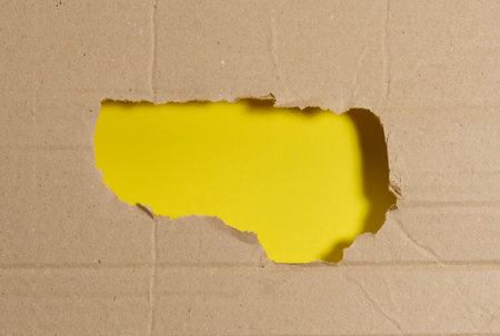 A rough, jagged hole in a sheet of brown corrugated cardboard, revealing a bright yellow background....