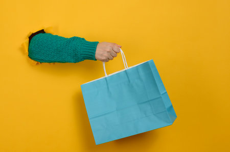 A woman's hand, clad in a green knitted sweater, holds a blue paper bag. The hand protrudes from a torn hole in a bright yellow background.の写真素材
