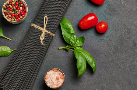 Black spaghetti, sprig of fresh basil, cherry tomatoes, pepper mix and salt on black table, top viewの写真素材