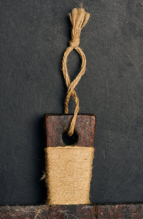 An old cutting board handle made of dark wood, wrapped in natural jute twine, with a loop at the top. It rests on a black textured background.の写真素材