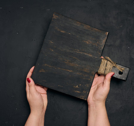A woman's hand holds the handle of an old, black wooden kitchen cutting board wrapped in twine against a black textured background. Recipe mockup.の写真素材