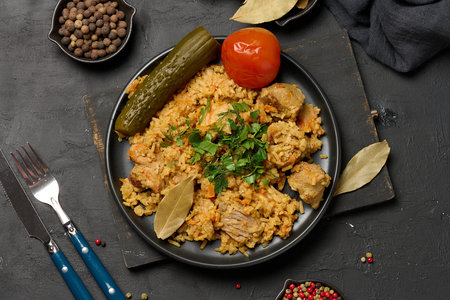Aromatic pilaf with pieces of meat and herbs served on a black plate with pickled cucumber,...