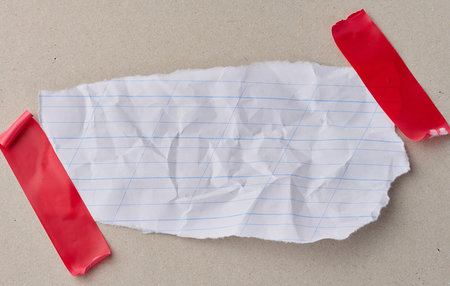 A scrap of white notebook paper with diagonal lines, attached with red electrical tape to gray cardboard. Copy space