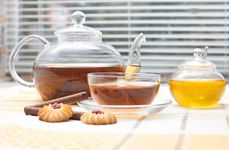 Glass teacup,  mug, honey jar and cakes on table near window with jalousieの写真素材