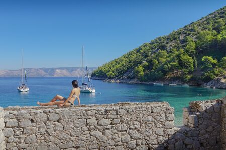 back view of a woman sits on the rocky wall with the view of turquoise sea water, two yachts, blue sky and green hills as a backgroundのeditorial素材