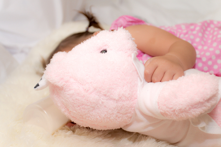 Baby drinking water or milk from a small bottle and lying on bed while hugging pink teddy bear in the bedroom because the baby having fever and attach cooling gel pad on her forehead for relief fever.の写真素材