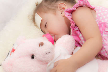 Baby drinking water or milk from a small bottle and lying on bed while hugging pink teddy bear in the bedroom because the baby having fever and attach cooling gel pad on her forehead for relief fever.の写真素材