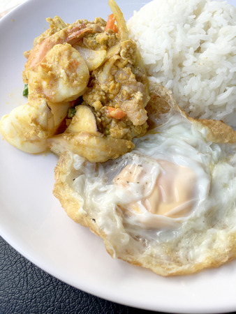 Stir fry prawns and squid with curry power thai style with fried egg with rice in white dish on black fabric texture background. Thai style food.の写真素材