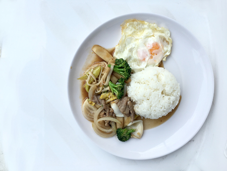 Thai cuisine. Stir-fried colorful vegetables, onion, broccoli, mushroom with meat in white plate with rice and fried egg on white background. Thai style food in fast street restaurant.の写真素材