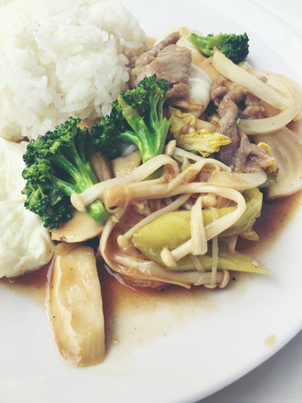 Thai cuisine. Stir-fried colorful vegetables, onion, broccoli, mushroom with meat in white plate with rice and fried egg on white background. Thai style food in fast street restaurant.の写真素材