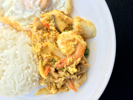 Stir fry prawns and squid with curry power thai style with fried egg with rice in white dish on black fabric texture background. Thai style food.の写真素材